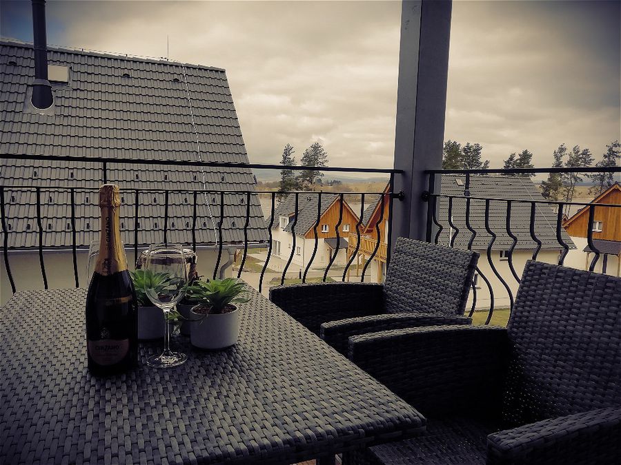A spacious balcony is furnished with a round table and two wicker chairs. A bottle of sparkling wine and two glasses are arranged on the table, accompanied by potted plants. The view reveals neighboring buildings and a cloudy sky in the background.