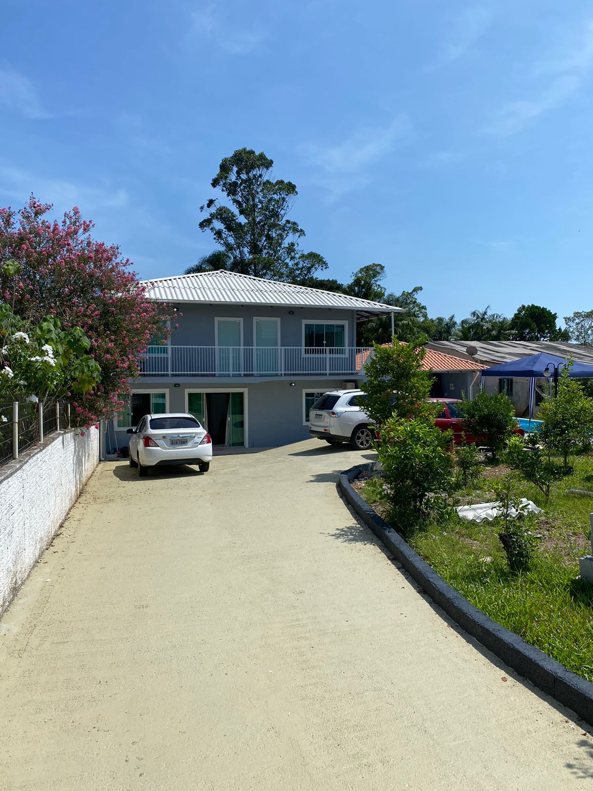 A two-story house is positioned at the end of a sandy driveway, surrounded by greenery. Two vehicles are parked in front, with flowering bushes and small trees visible in the landscaped yard. Clear blue skies are seen above, contributing to a bright ambiance.