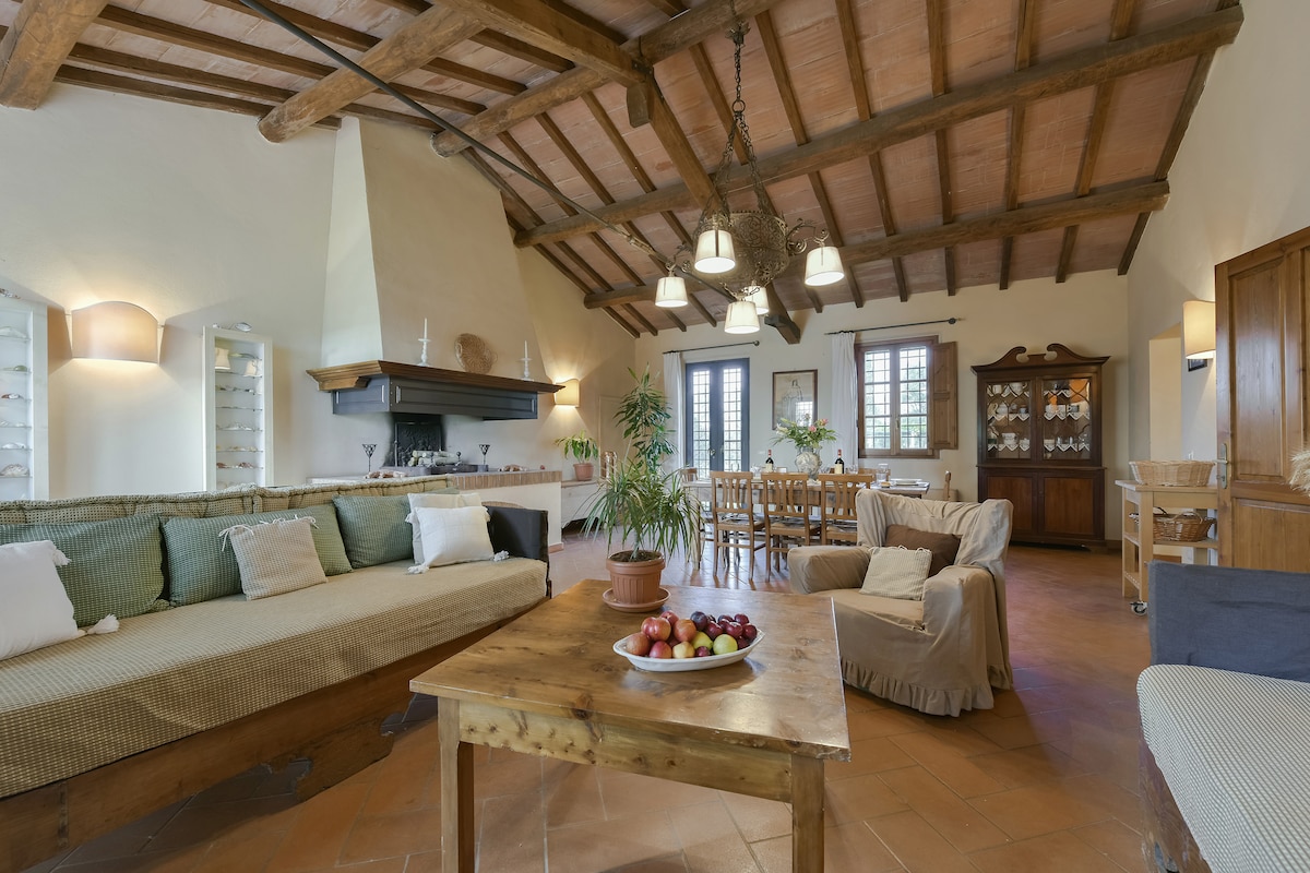 A spacious living area features high wooden beams and warm terracotta flooring. A cozy sofa, adorned with cushions, invites relaxation, while a wooden coffee table displays a bowl of fresh fruits. A dining area with a large table is visible, illuminated by natural light from the windows.