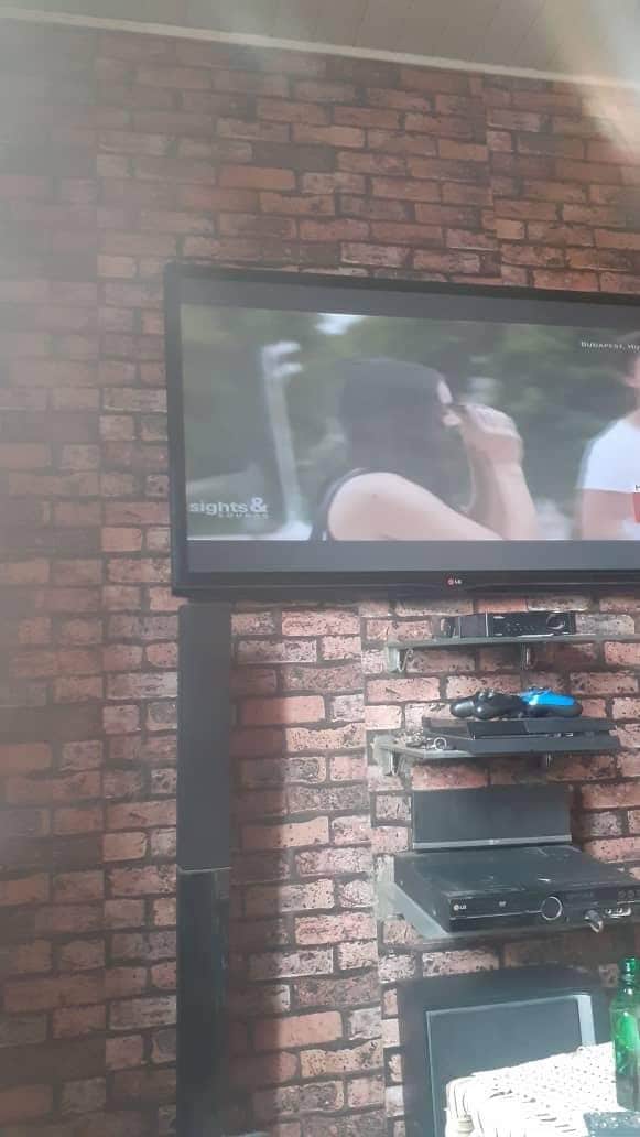 A wall-mounted television is positioned above a black entertainment center against a brick-patterned wall. The entertainment center holds various electronic devices. Soft light creates a warm ambiance within the compact living space.