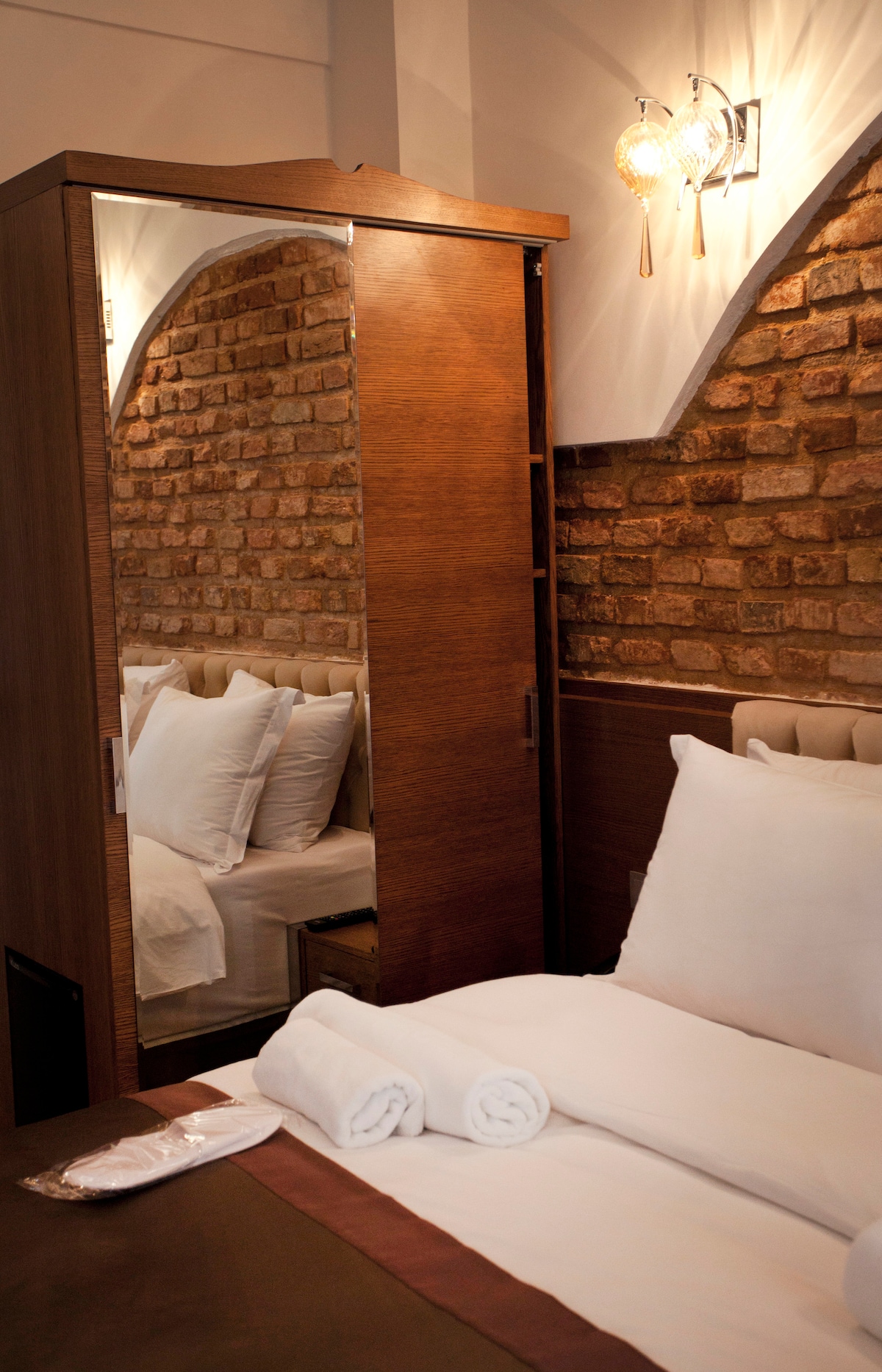 A cozy bedroom is featured, showcasing a mirrored wardrobe with wooden detailing against a warm brick wall. A neatly made bed with white pillows and a rolled towel is presented, providing a clean and inviting space.