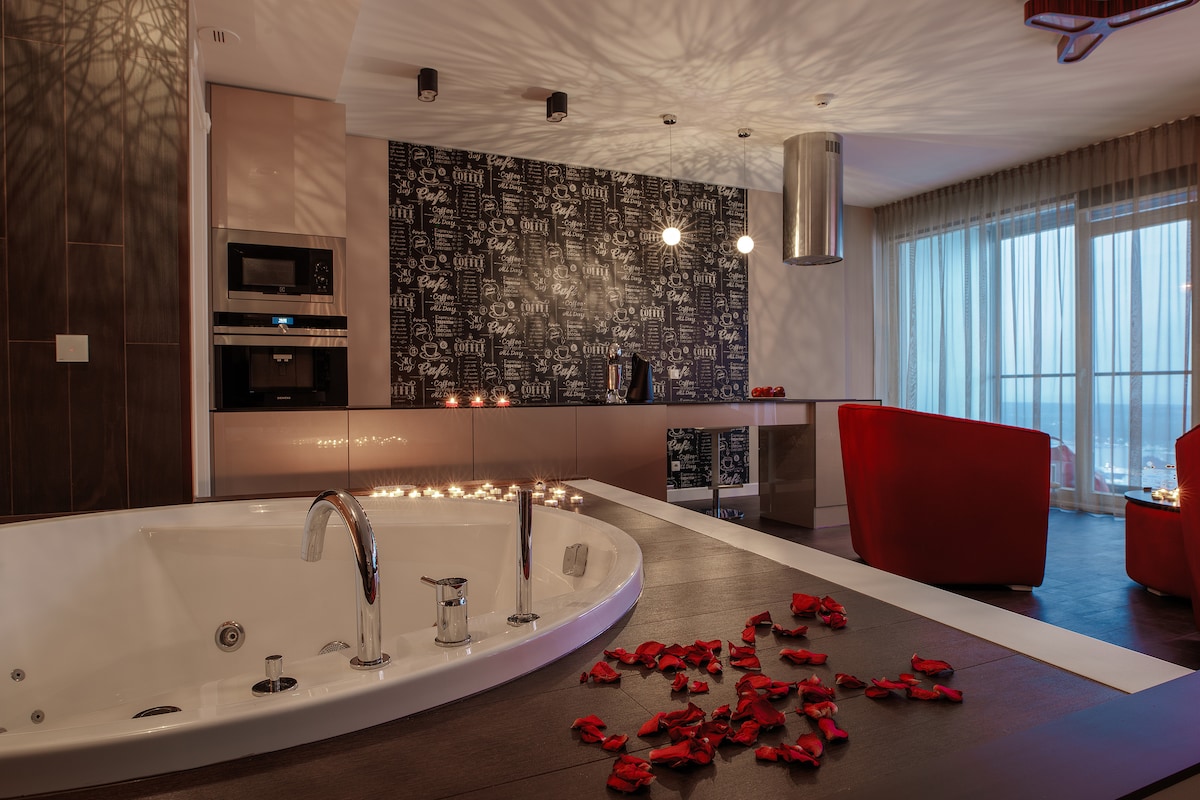 A spacious jetted bathtub is positioned near a modern kitchen featuring high-quality appliances. The wall behind is adorned with a decorative pattern, while soft lighting creates a warm ambiance. Red rose petals scatter over the dark wood floor, complementing the stylish red seating area.