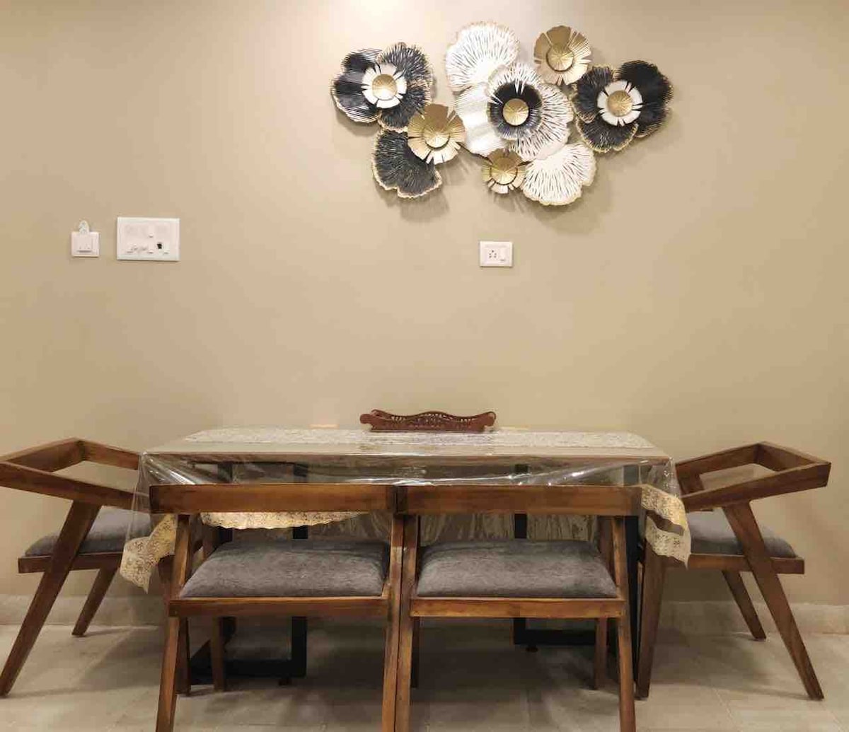 A dining area is presented with a wooden table and four elegant chairs, each featuring soft cushioned seats. A decorative wall piece with floral designs adds a unique touch above the table. The setting offers a warm and inviting space for shared meals.