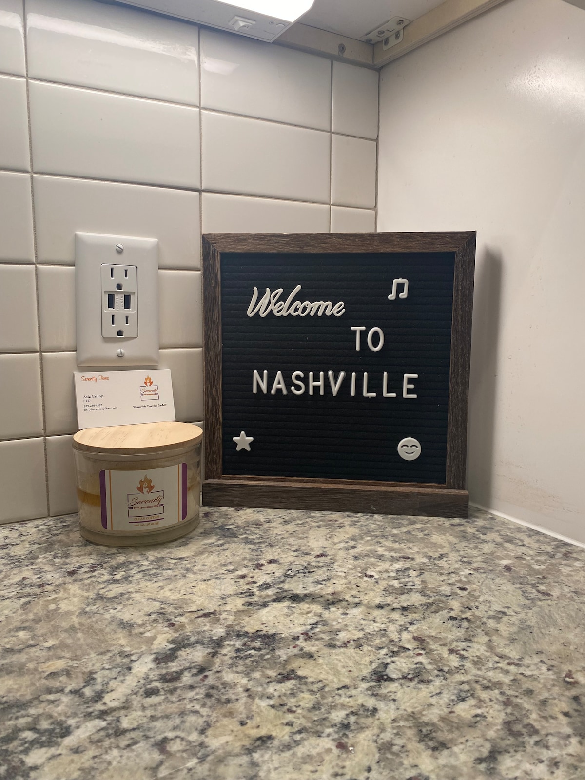 A warm welcome is extended with a decorative sign that reads 'Welcome to Nashville,' positioned on a granite countertop. Nearby, a candle adds a cozy touch, alongside a small business card on display. The surroundings are characterized by clean white tile walls and modern electrical outlets.