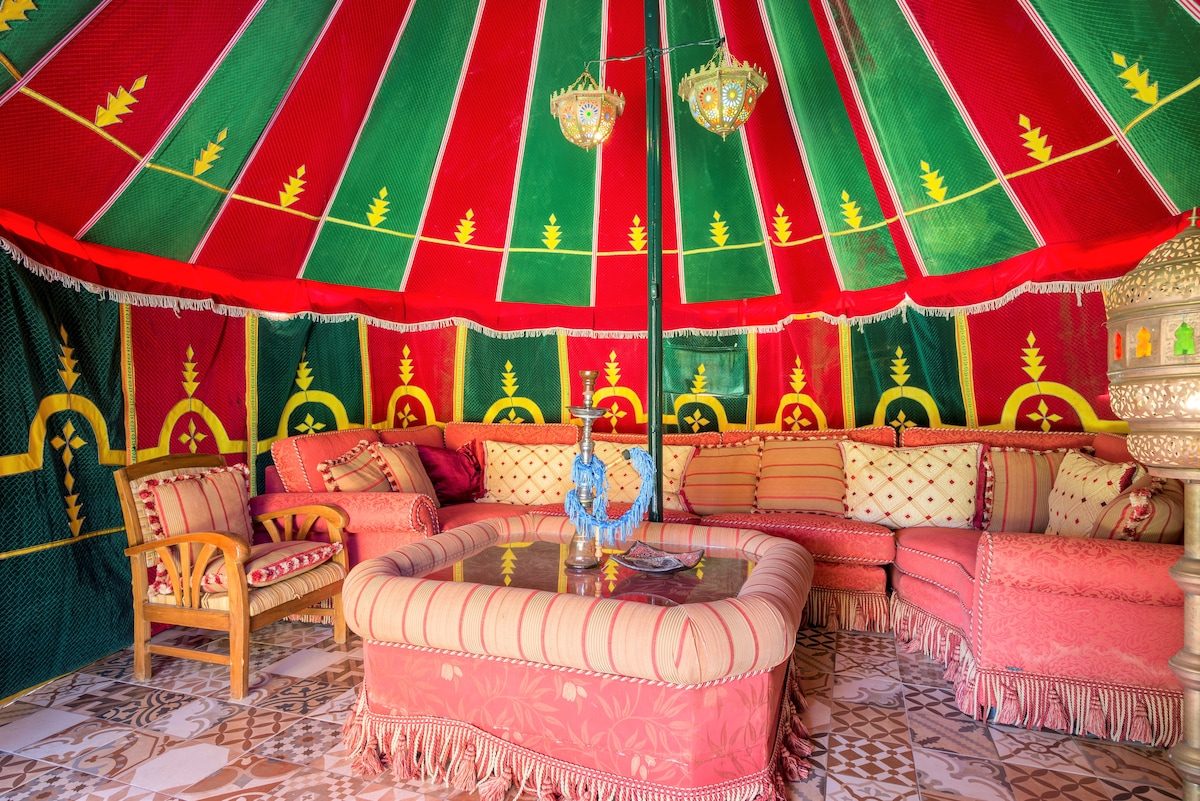 A vibrant Moroccan Haima is featured, adorned with bold red and green fabric. Comfortable seating is arranged around a central table, inviting relaxation. Decorative lighting elements hang from the ceiling, providing a cozy atmosphere within the intricately patterned tiled space.