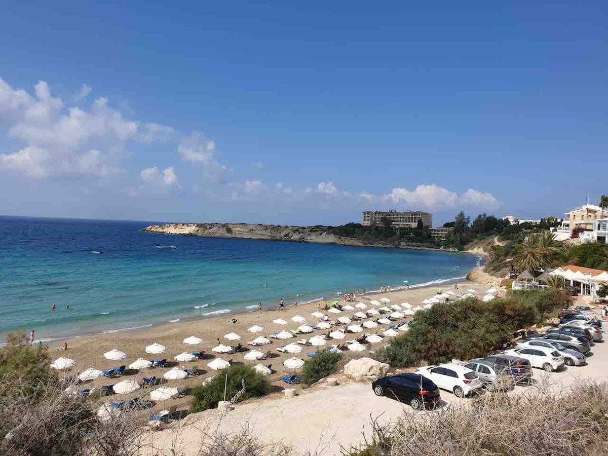 A panoramic view of a sandy beach is presented, featuring numerous sunbeds and umbrellas arranged neatly across the shore. The tranquil turquoise water meets the coastline, and distant hills provide a natural backdrop, while a few vehicles are parked along the road nearby.