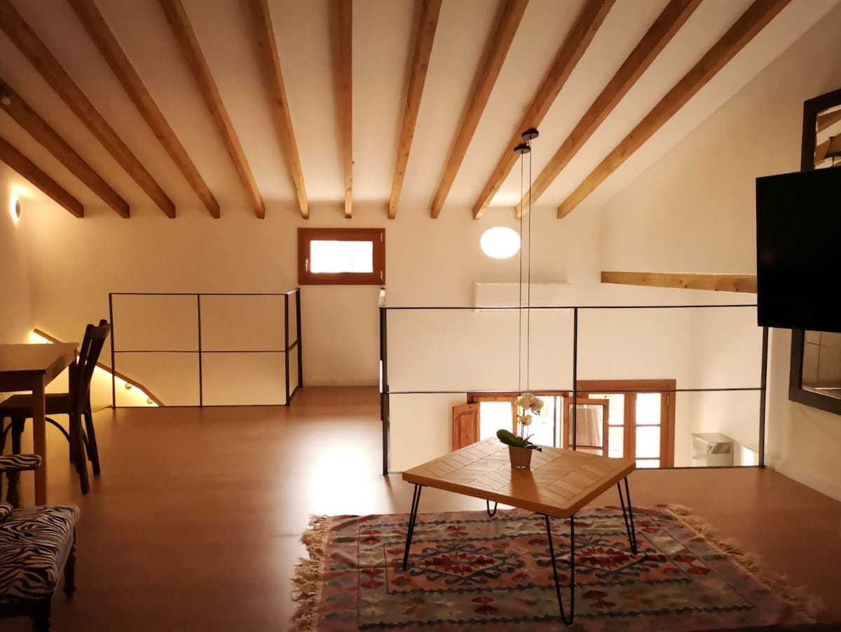 An upper deck living area features a wooden ceiling with exposed beams, enhancing the open space. A low wooden table is positioned on a colorful rug, complementing the light tones of the room. Natural light enters through a window, illuminating the inviting atmosphere.