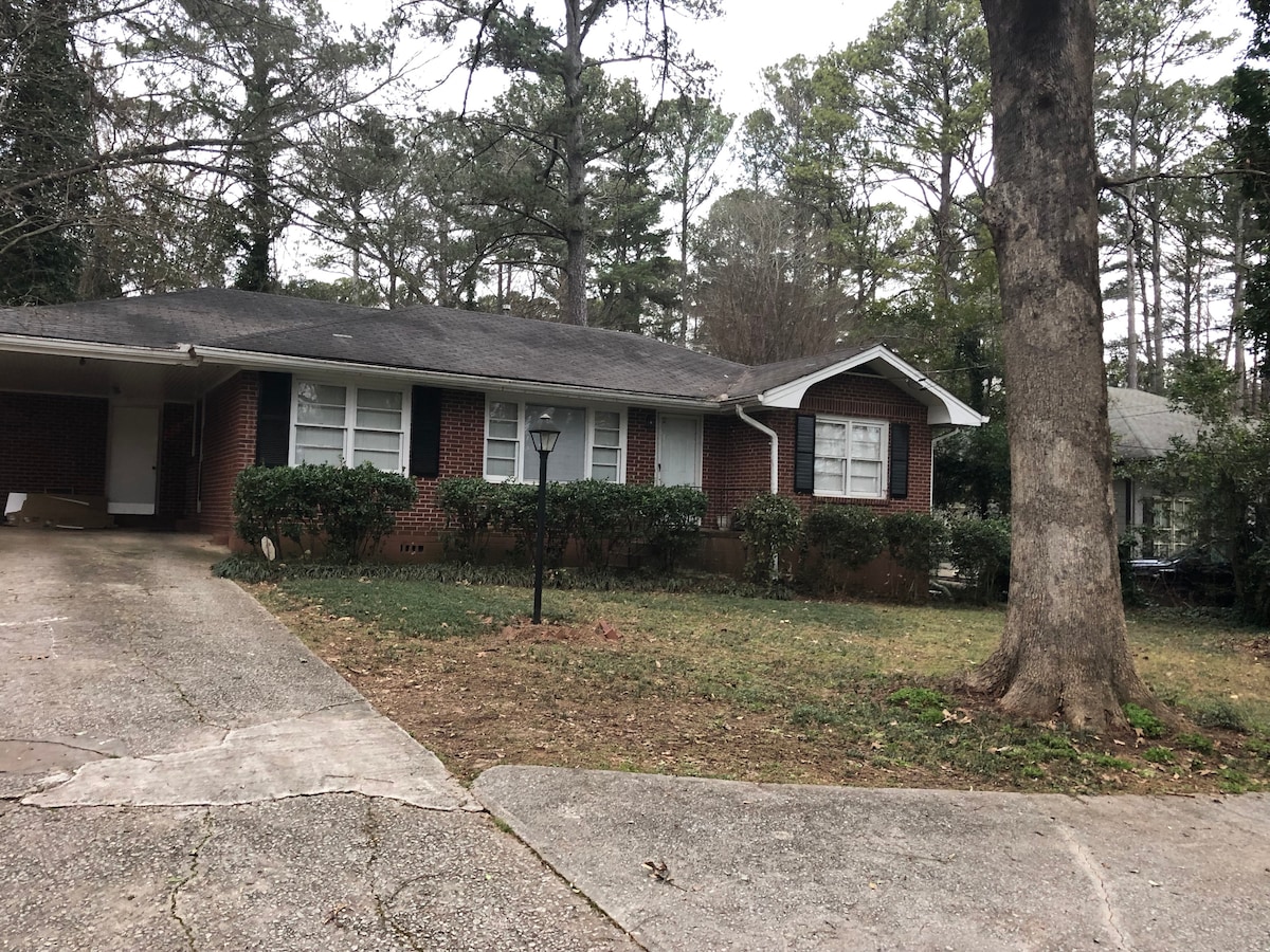 A single-story brick house is surrounded by towering trees and a manicured lawn. The home features a covered carport and multiple windows, allowing natural light to fill the interior. A pathway leads to the entrance, offering easy access to the front door.