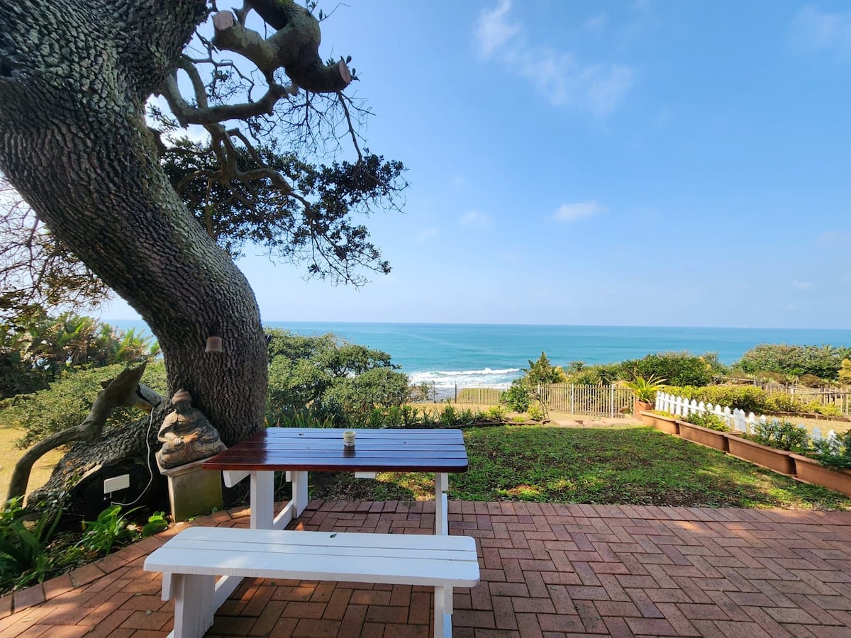 A shaded outdoor seating area features a wooden table with benches, positioned on a brick patio. The view stretches towards the ocean, where the water meets the sky. Lush greenery and a sandy beach are visible in the background, enhancing the serene coastal setting.