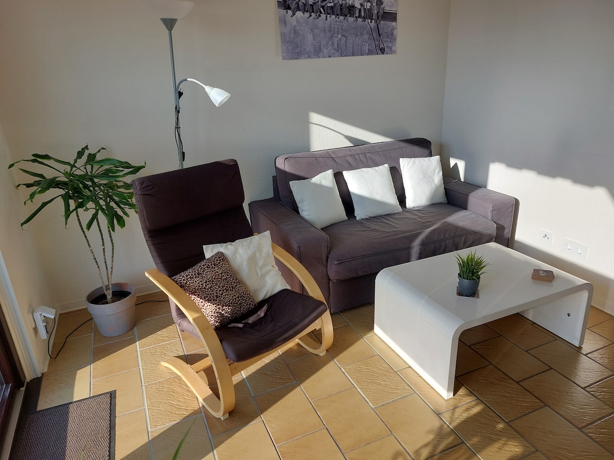 A cozy seating area features a dark sofa accompanied by white pillows and a modern armchair. A small white coffee table rests on the tiled floor, complemented by a decorative potted plant. Natural light filters in, highlighting the inviting arrangement.