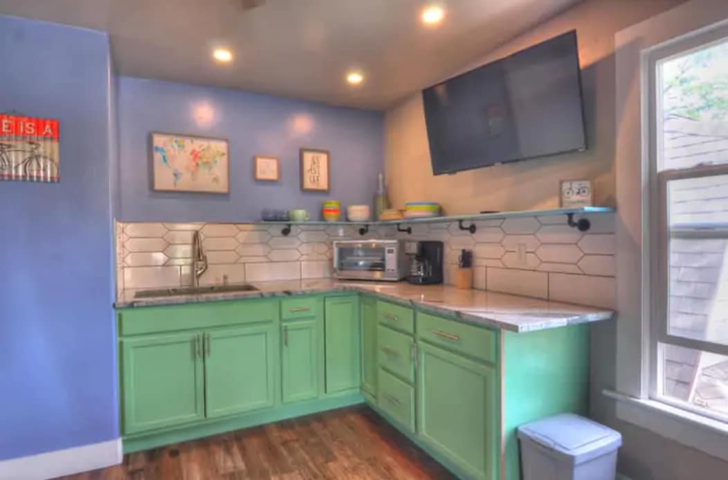 The kitchenette features green cabinetry with a modern design, complemented by a light-colored countertop. Various kitchen appliances, including a microwave and coffee maker, are visible. A wall-mounted television and a window allow for natural light, enhancing the functional space.