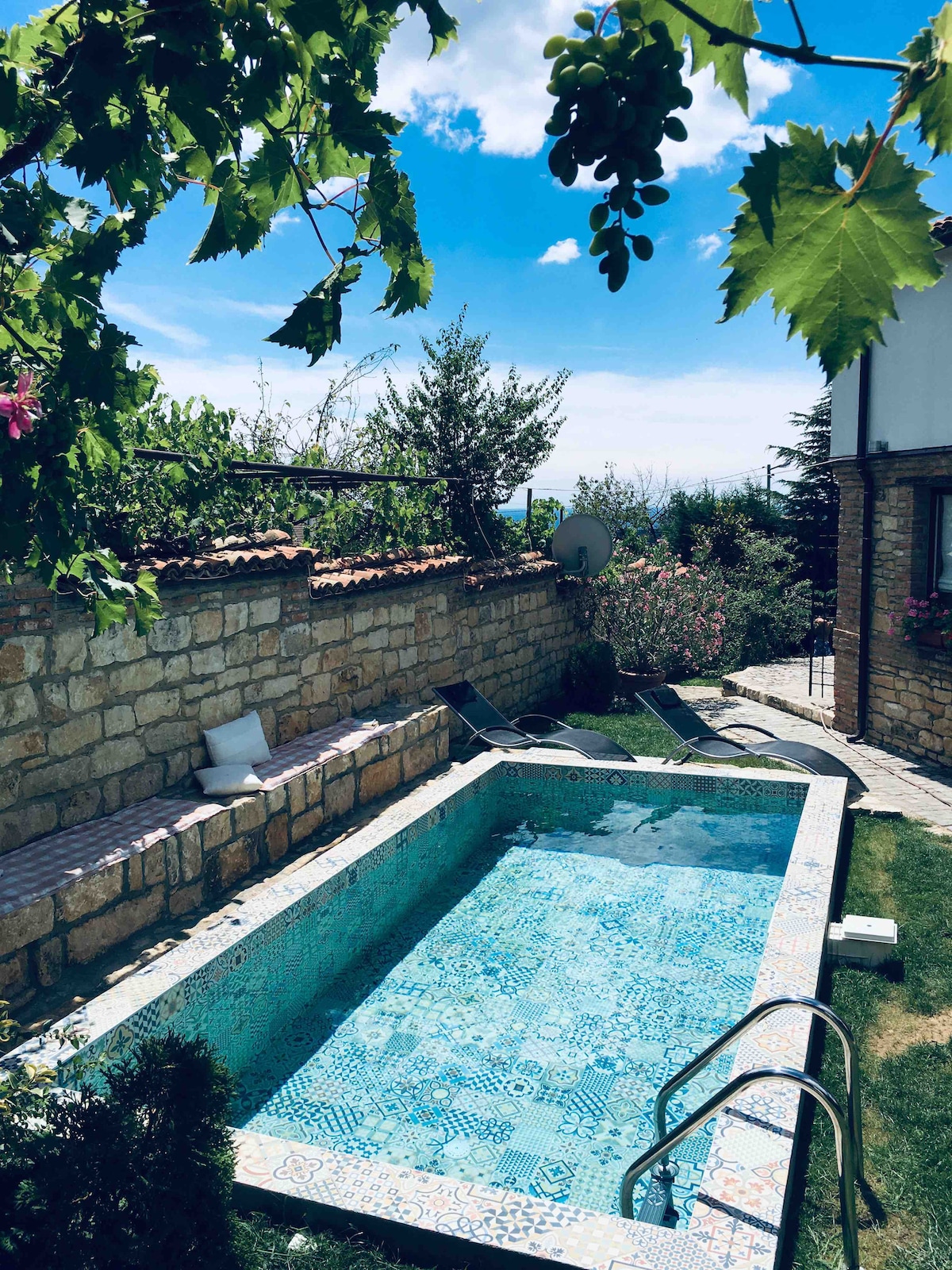 A serene swimming pool is framed by vibrant greenery and stone walls. Comfortable lounge chairs are positioned nearby, inviting relaxation. The crystal-clear water reflects the sky, while a well-maintained garden adds to the tranquil ambiance.
