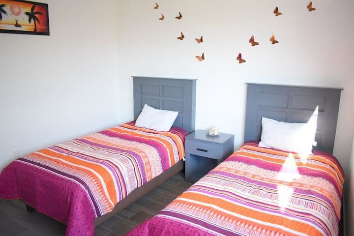 A bright bedroom features two twin beds with colorful striped blankets. Each bed is positioned beside a small nightstand. Butterfly decals adorn the walls above, complementing the light-colored space and enhancing the room's cheerful ambiance.