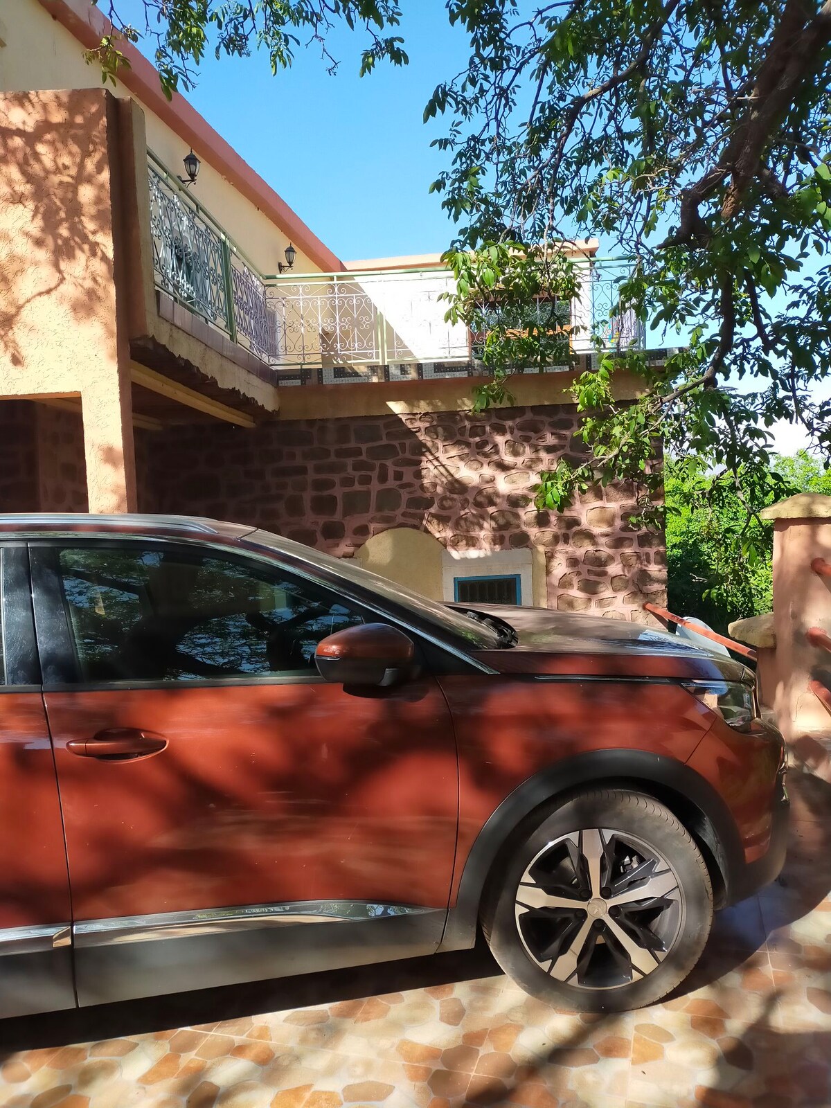 A car is parked on a patterned stone surface, with sunlight filtering through surrounding trees and casting playful shadows. The building, constructed with a combination of stone and earth, features a balcony above and a well-maintained exterior.