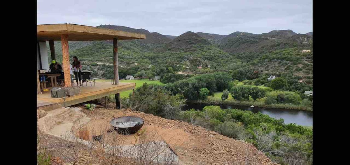 A wooden deck extends from the cottage, offering views of lush greenery and rolling hills. A fire pit is positioned on the ground below, with the Goukou river winding through the landscape. The tranquil surroundings invite exploration of nature.