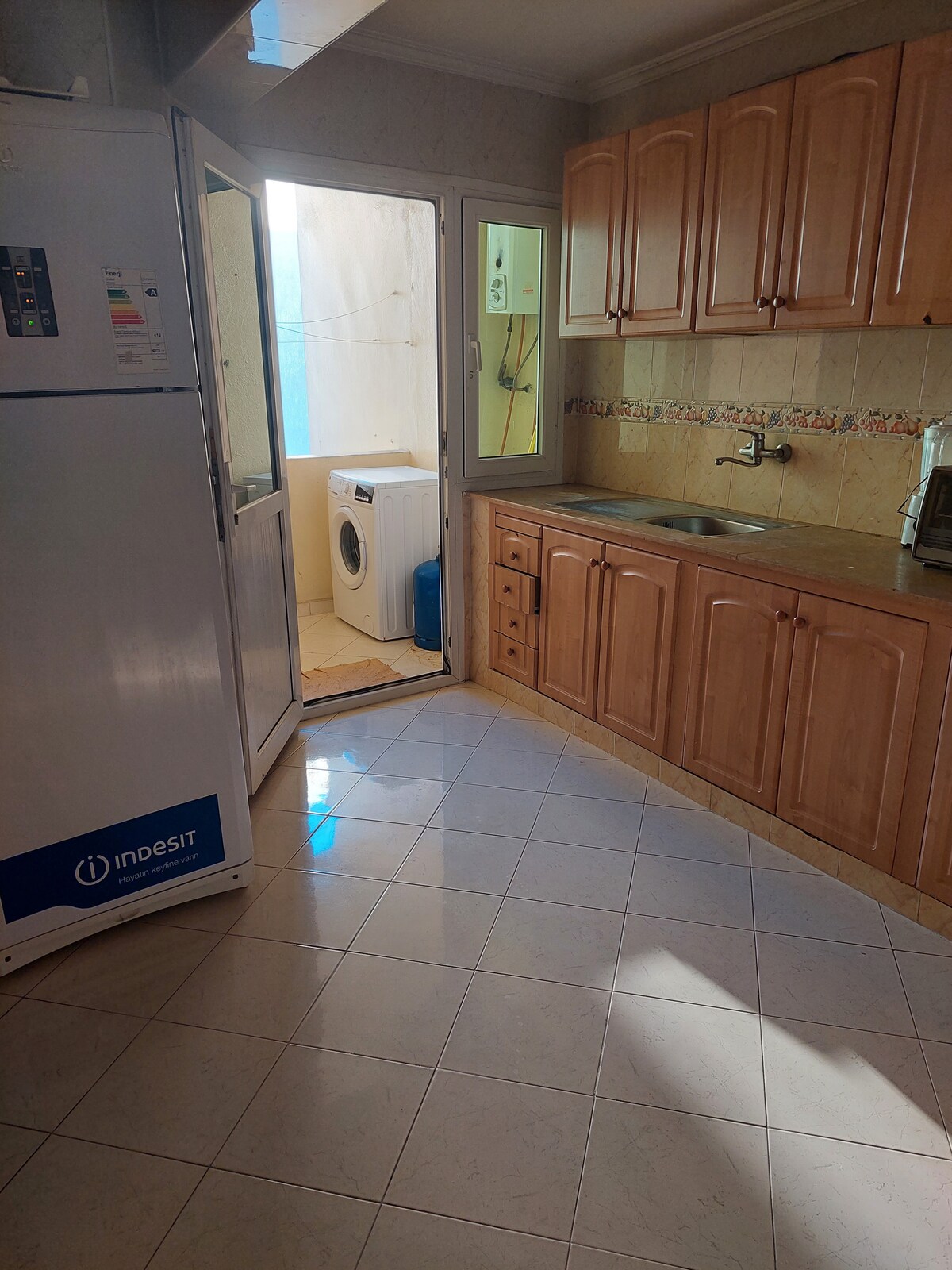 The kitchen features wooden cabinetry and a tiled floor, illuminated by natural light. A white refrigerator and a washing machine are present. A sink is situated under the window, with an adjacent door leading to an outdoor area.