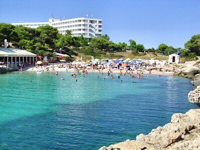 A sandy beach is highlighted by clear turquoise waters, surrounded by gentle waves. Beach umbrellas and sunbathers occupy the shore, while nearby, a beach bar contributes to a lively atmosphere. The distant background features a hotel set amidst lush greenery.