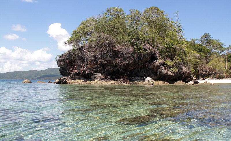 A rocky shoreline is visible with lush greenery lining the edges, contrasting against the clear, shallow waters. Sunlight reflects off the water's surface, creating a serene and tranquil ambiance, while distant hills fade into the horizon beneath a blue sky with scattered clouds.