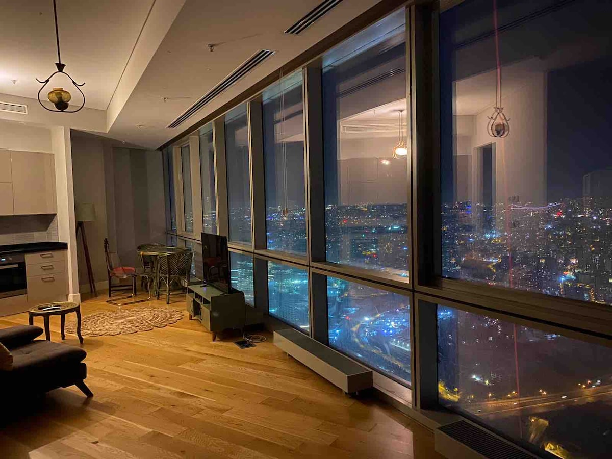 A modern living area features large floor-to-ceiling windows, providing expansive views of the city lights at night. A small television is positioned on a stand across from a comfortable seating area. A round rug is placed on the wooden floor, enhancing the cozy atmosphere.