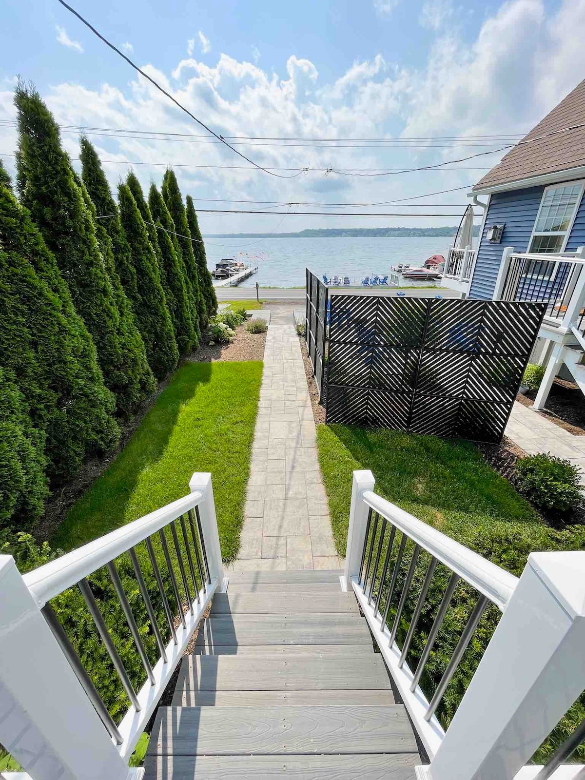 A well-maintained set of stairs leads down from the cottage to a lush green lawn, with tall evergreen trees providing a natural border. The pathway is flanked by neat landscaping, and the serene lake is visible in the distance under a blue sky.