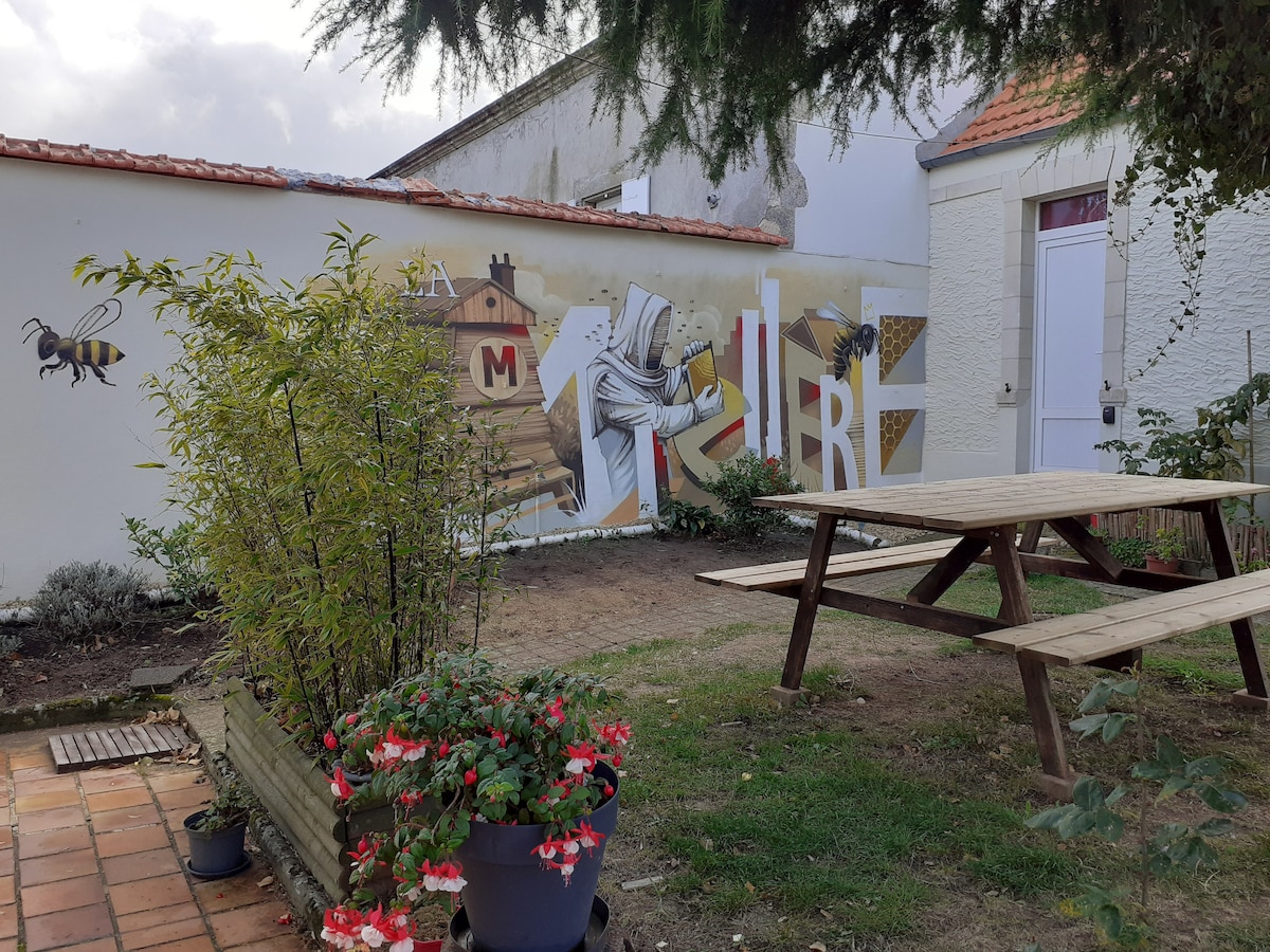 An outdoor space is featured with a mural depicting beekeeping, surrounded by greenery and flower pots. A wooden picnic table sits on the lawn, complemented by a neat garden bed. The area is enclosed, providing a private setting for relaxation.