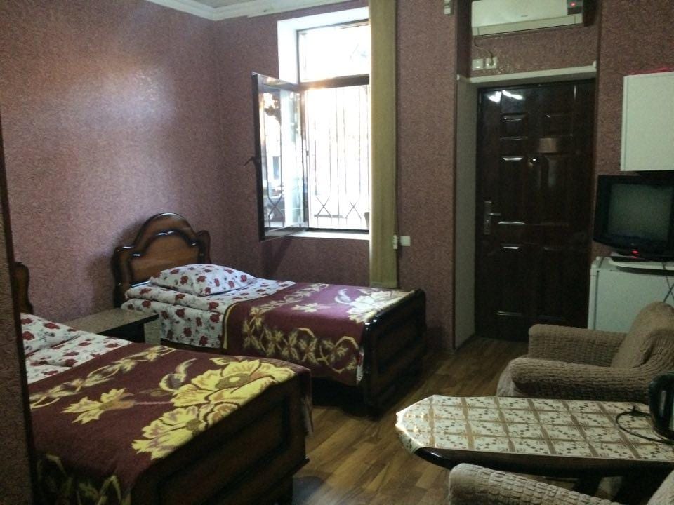 A room features two beds with floral-patterned covers, positioned adjacent to a window that opens to the outside. A wooden table is placed between the beds, and a cozy seating area is provided with a woven chair. A television and door are visible along the wall.
