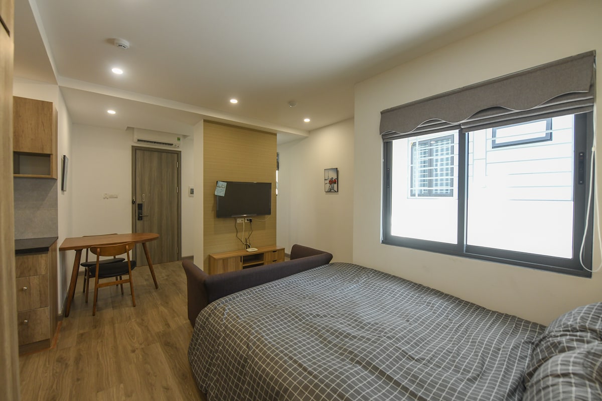 A well-furnished room features a spacious bed covered with a checkered blanket. A compact dining area is situated near a window that invites natural light. A television is mounted on the wall opposite the bed, contributing to a functional living space.