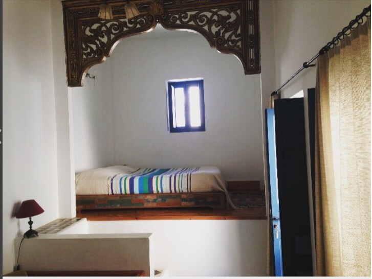 A cozy sleeping area is elevated within a traditional wooden alcove, featuring a spacious bed adorned with a striped blanket. A small lamp sits on a nearby table. Light enters through a window with wooden shutters, while curtains softly frame the space.