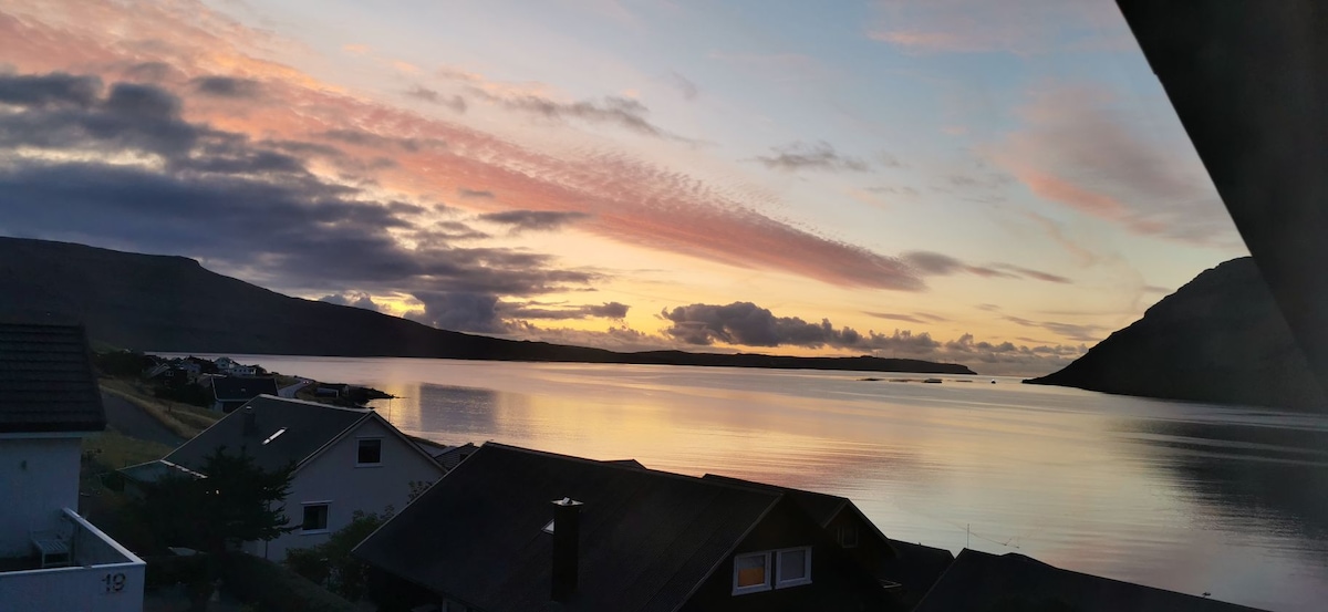 A serene landscape captures a tranquil body of water reflecting soft pastel colors of a sunset. Nearby houses are visible, nestled along the shore, with a backdrop of hills gently meeting the sky in the distance.