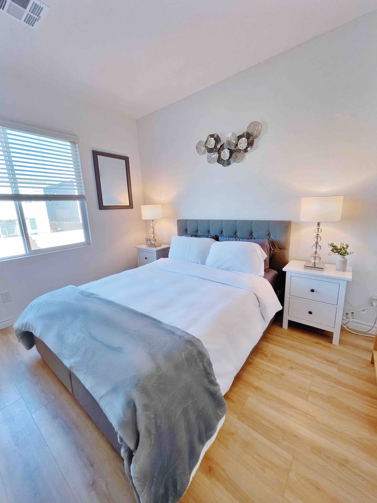 A modern bedroom features a cozy queen bed dressed in crisp white linens, complemented by a soft grey throw. Two bedside tables with lamps are positioned next to the bed. Natural light fills the space through a window adorned with wooden blinds, and decorative wall art enhances the ambiance.