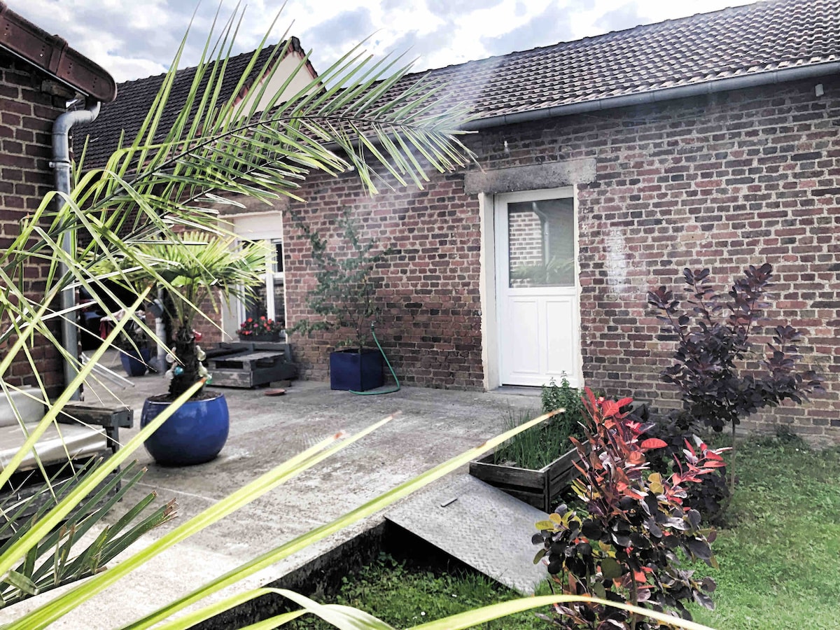 A serene outdoor space features a concrete patio surrounded by low greenery and potted plants. A white door leads to the interior, while the brick exterior of the building provides rustic charm. Sunlight filters through nearby palm leaves.