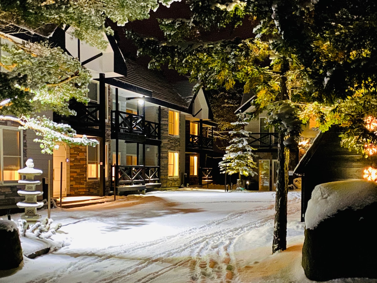 A serene winter scene captures the exterior of the property, adorned with a fresh layer of snow. Soft lighting illuminates the building's architectural details, while trees and stone features contribute to a tranquil atmosphere.