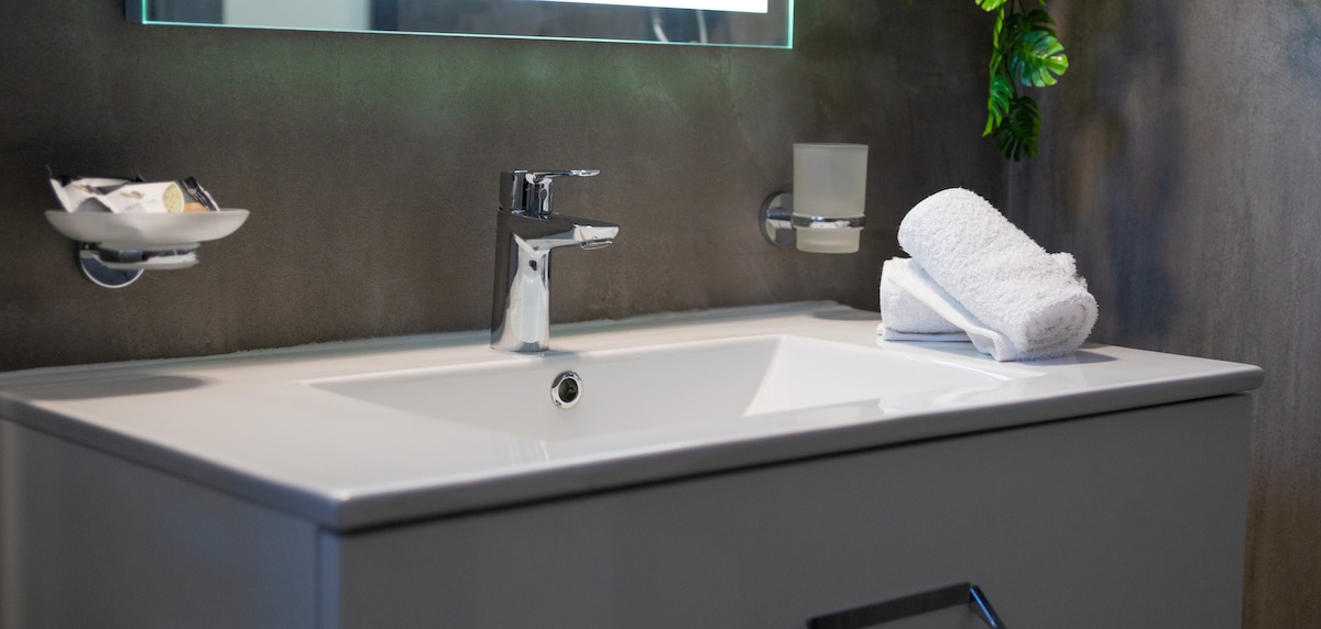 A modern bathroom sink area features a sleek, minimalist design. A large mirror hangs above the basin. White towels are neatly arranged beside the faucet, providing a clean and functional appearance. Subtle greenery is included in the corner, adding a touch of nature.