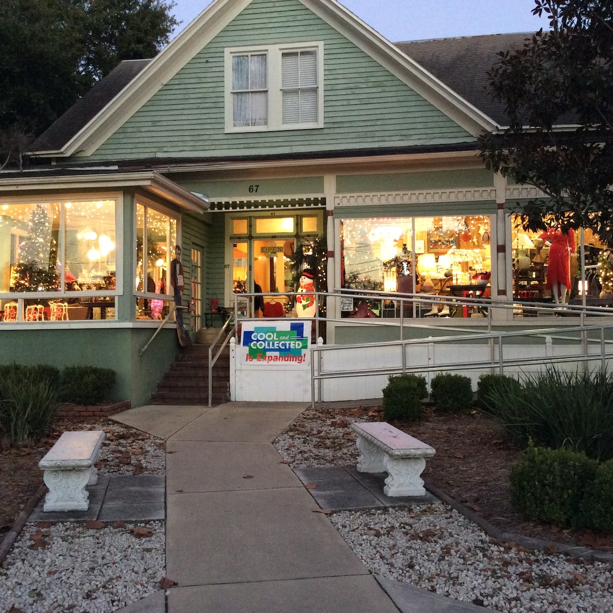 A charming vintage store is shown in soft evening light. The green exterior features large windows displaying colorful merchandise. Two white benches are positioned on a pathway lined with shrubs, leading to a welcoming entrance adorned with holiday decorations.