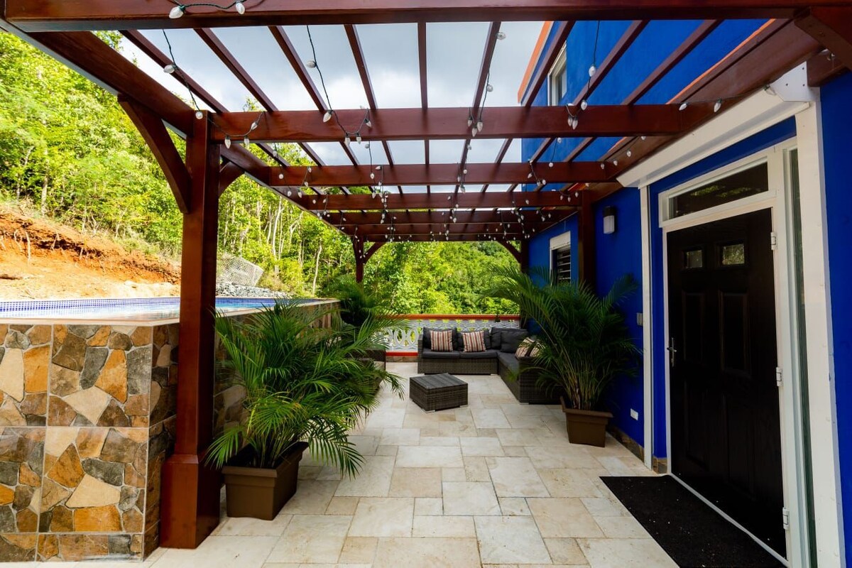 An inviting outdoor patio area features a wooden pergola adorned with string lights. Comfortable seating arrangements include a low table surrounded by potted plants, with a stone wall and a glimpse of the pool visible in the background.