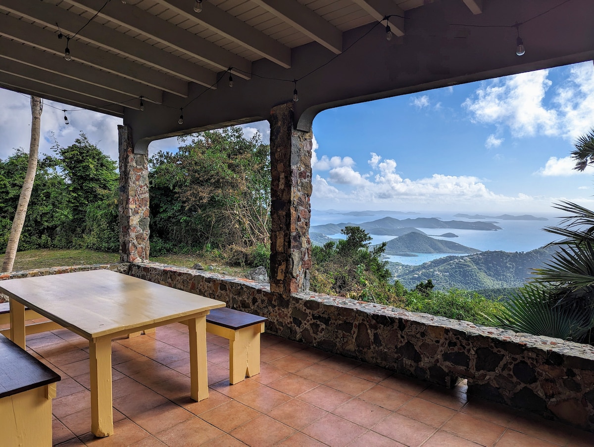 An outdoor dining area is framed by natural stone columns, with a large yellow table and wooden benches accommodating several guests. A panoramic view of the ocean and distant islands is showcased through wide openings, complemented by a clear sky and lush greenery.