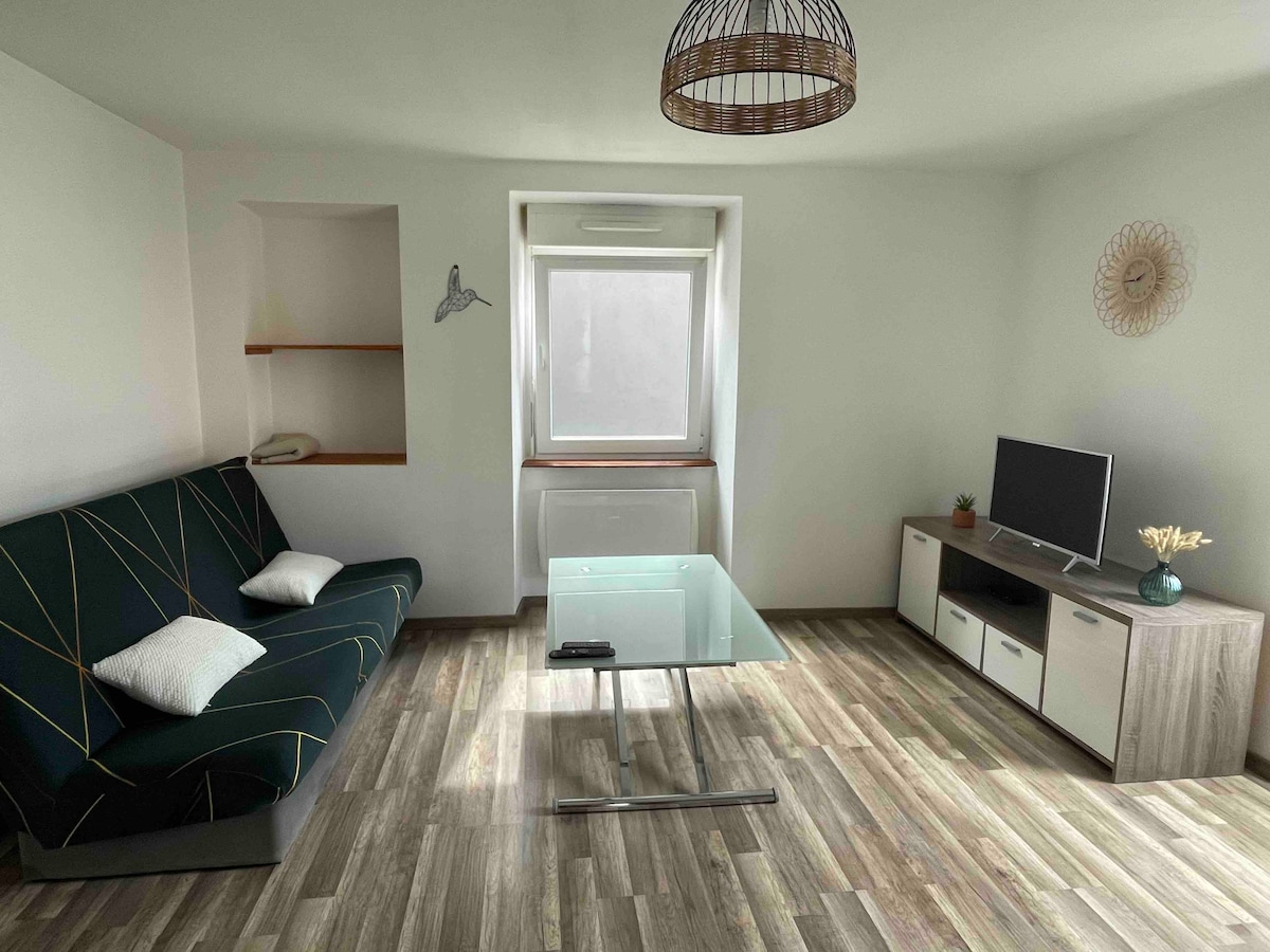 A comfortable living area features a dark green sofa with geometric patterns and two white cushions. A glass coffee table sits centrally, complemented by a low wooden entertainment unit, which holds a television. Natural light enters through a large window, enhancing the space.