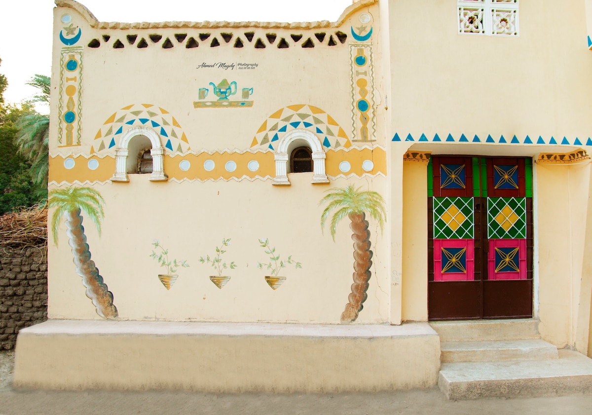 A portion of a building is showcased, featuring vibrant murals inspired by Nubian culture. Two arched windows with colorful designs are present, flanked by decorative palm tree motifs. The main entrance is adorned with intricate geometric patterns in bright colors on the wooden doors, set against a light-colored wall.