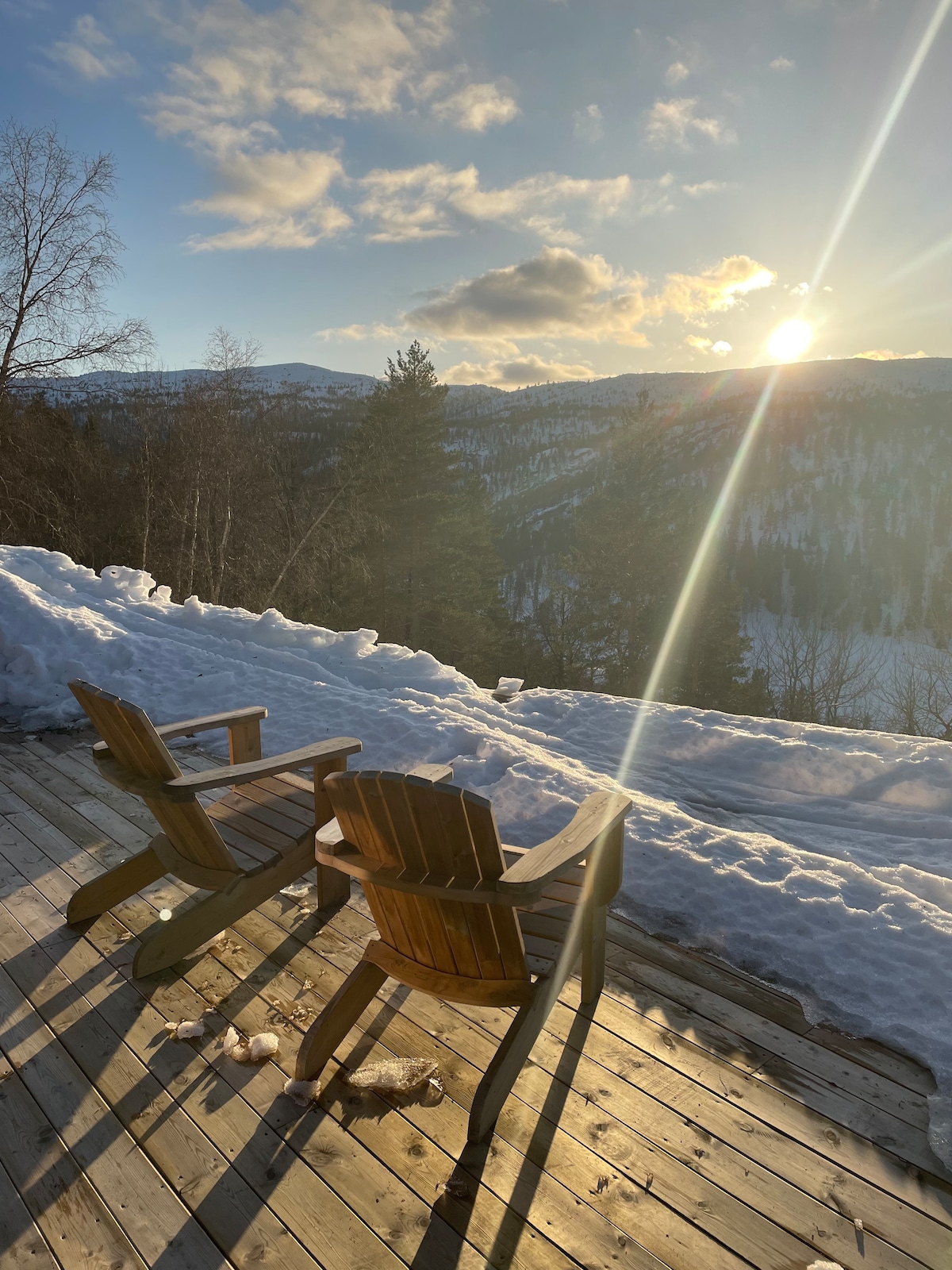 Two wooden chairs are positioned on a deck overlooking a snow-covered landscape. The sun sets behind distant mountains, casting golden light across the scene. Snow blankets the ground, while trees line the view in the background.