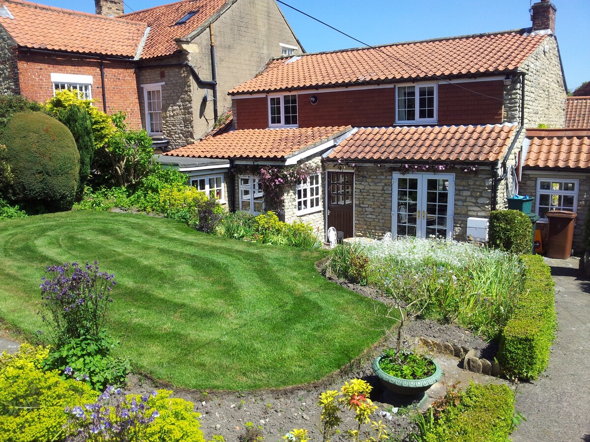 A well-maintained garden features neatly trimmed grass, bordered by colorful flower beds. The cottage's rustic stone façade and red-tiled roof complement the lush greenery, with multiple windows allowing natural light to enhance the inviting exterior. A path leads towards the entrance of the cottage.