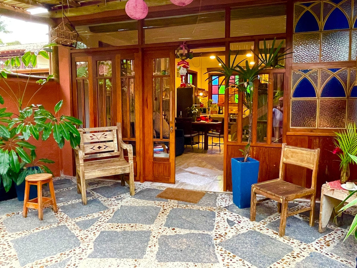 A welcoming entryway features a stone pathway leading to wooden doors with large glass panels. The area includes two simple wooden chairs and a small stool surrounded by potted plants, creating a harmonious connection to nature.