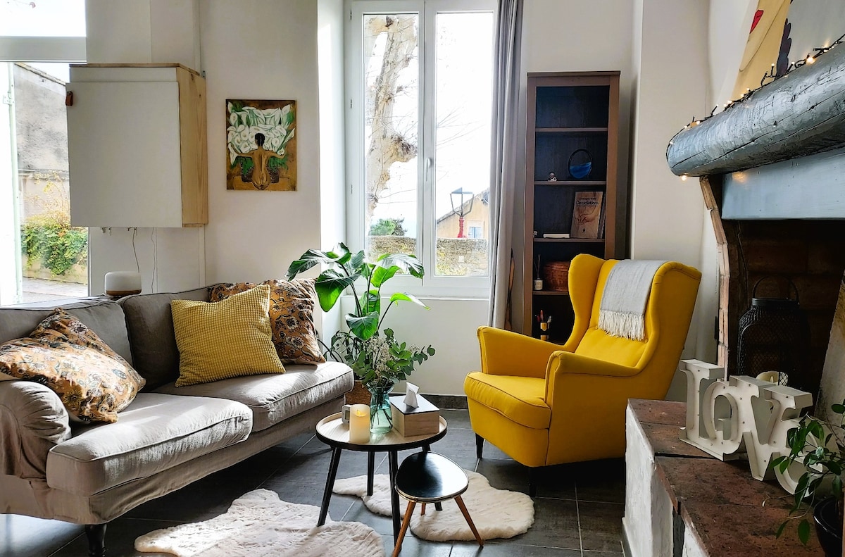 The living area features a light gray sofa paired with a bright yellow armchair, inviting relaxation. A round, dark wood coffee table is centered on a soft area rug. Large windows allow natural light to fill the space, complemented by decorative plants and artwork.