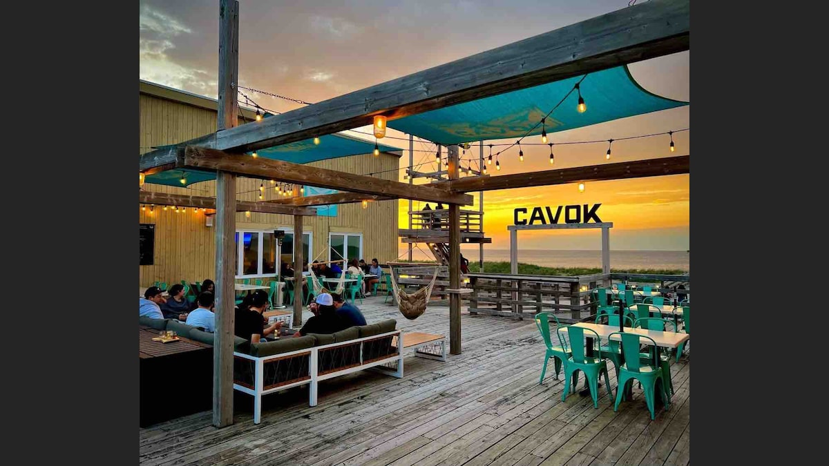 An outdoor deck area is highlighted, featuring string lights and shaded seating under a large canopy. Tables with teal chairs are arranged, and a fire pit area is visible. The beach and sunset create a serene backdrop, enhancing the inviting atmosphere.
