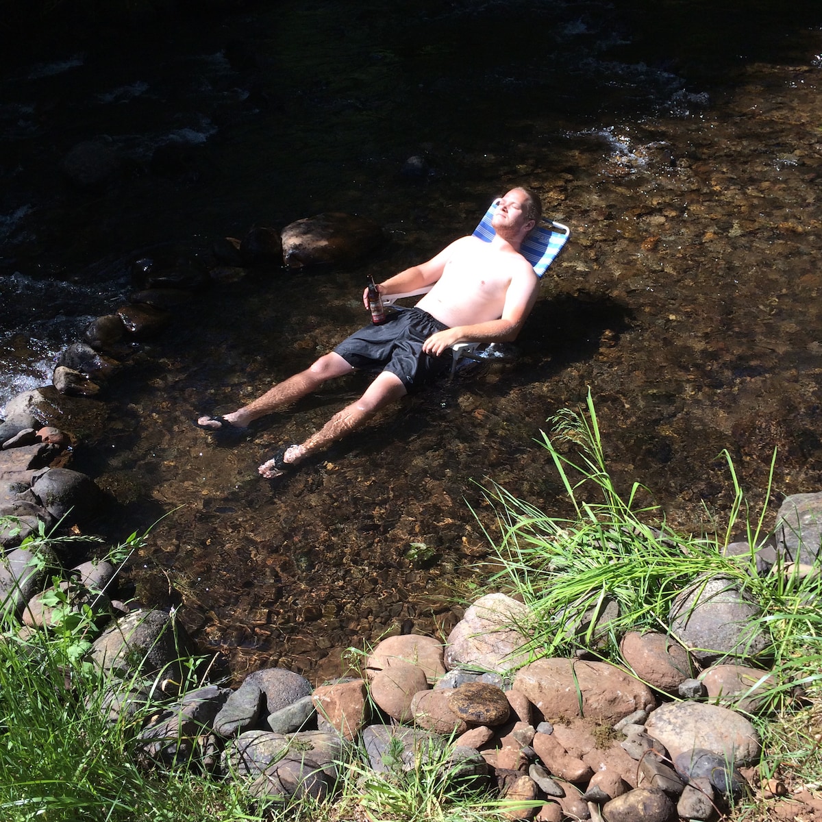 A person is seen reclining in a shallow, clear section of the Roaring River, surrounded by smooth stones and lush green grass. The gentle flow of the water creates a serene atmosphere, offering a peaceful spot for relaxation.