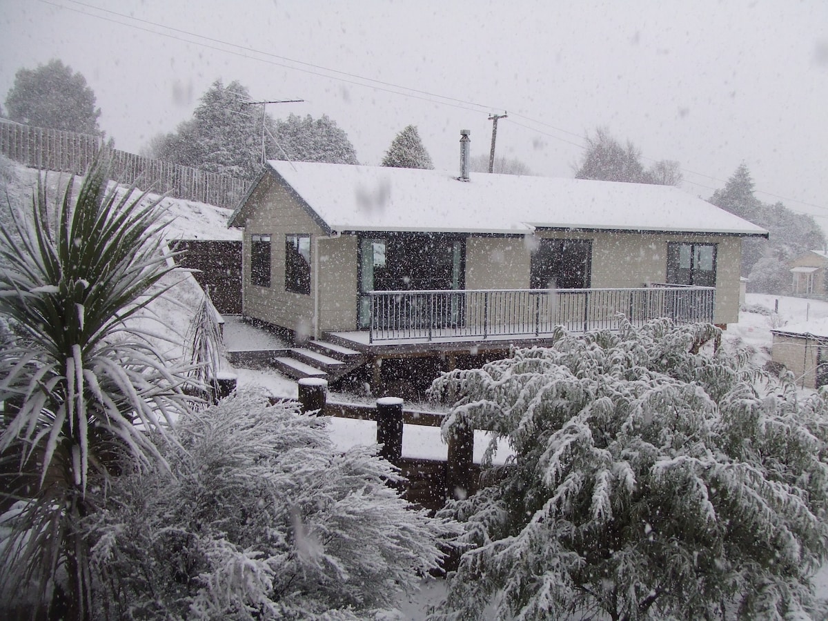 A snow-covered chalet is depicted, surrounded by white-dusted trees and shrubs. The building features a sloped roof and a front deck with railing. Snow falls steadily, creating a serene winter landscape in this alpine setting.