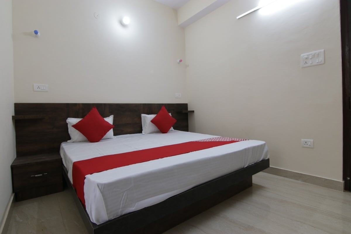 A well-furnished bedroom features a spacious bed with red accent pillows. The minimalistic design includes bedside tables and soft lighting, contributing to a calming environment. Light-colored walls and tiled flooring enhance the airy feel of the room.