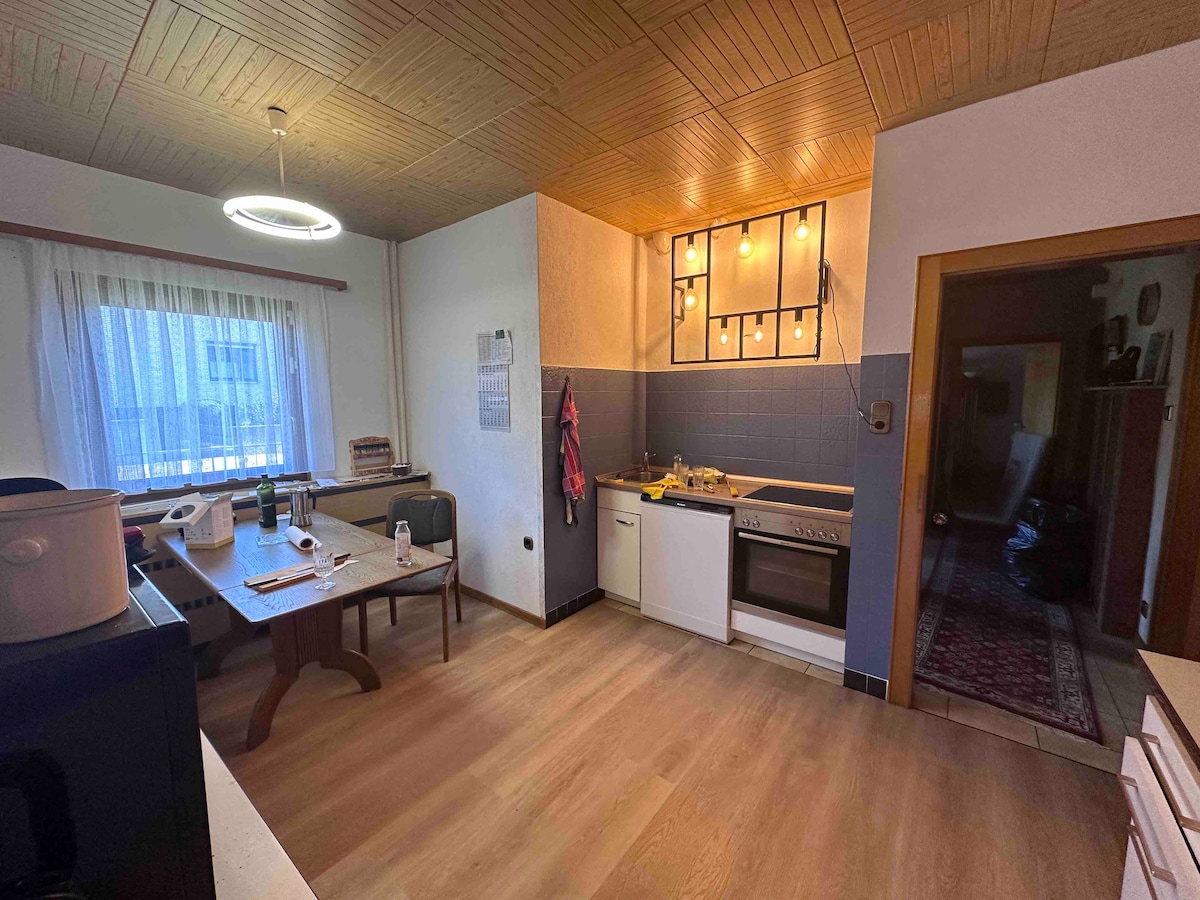 A functional kitchen is illuminated by pendant lights, featuring a wooden ceiling and ample countertop space. A dining table with four chairs is positioned nearby, while appliances include a stove, oven, refrigerator, and sink. A window provides natural light, with curtains partially drawn.