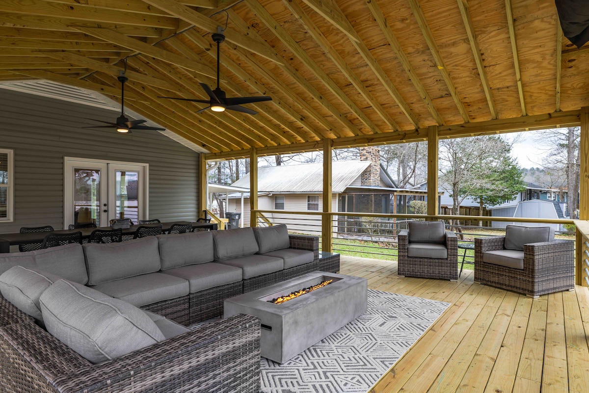 An oversized covered deck features a large sectional sofa and two chairs arranged around a concrete gas fire table. Two ceiling fans are visible above, and a patterned rug lies beneath the seating area, overlooking a scenic outdoor space.