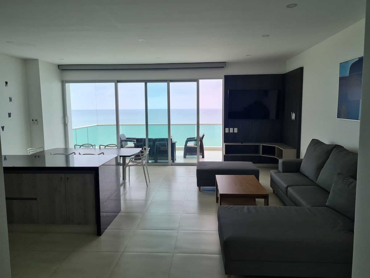 A modern living space features a comfortable sectional sofa and a wooden coffee table, facing a floor-to-ceiling window with a sweeping ocean view. A dining area with a glass table accommodates several chairs. The room is illuminated by natural light, offering a serene atmosphere.