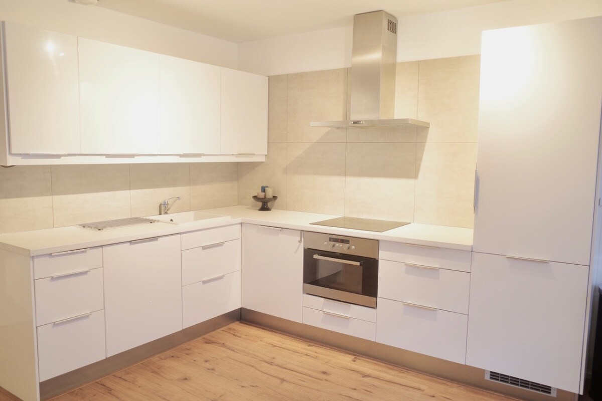 A modern kitchen space is designed with sleek white cabinetry and a neutral-toned backsplash. Stainless steel appliances, including an oven and extractor hood, are integrated into the layout. Ample counter space is available for meal preparation, complemented by warm wood flooring.